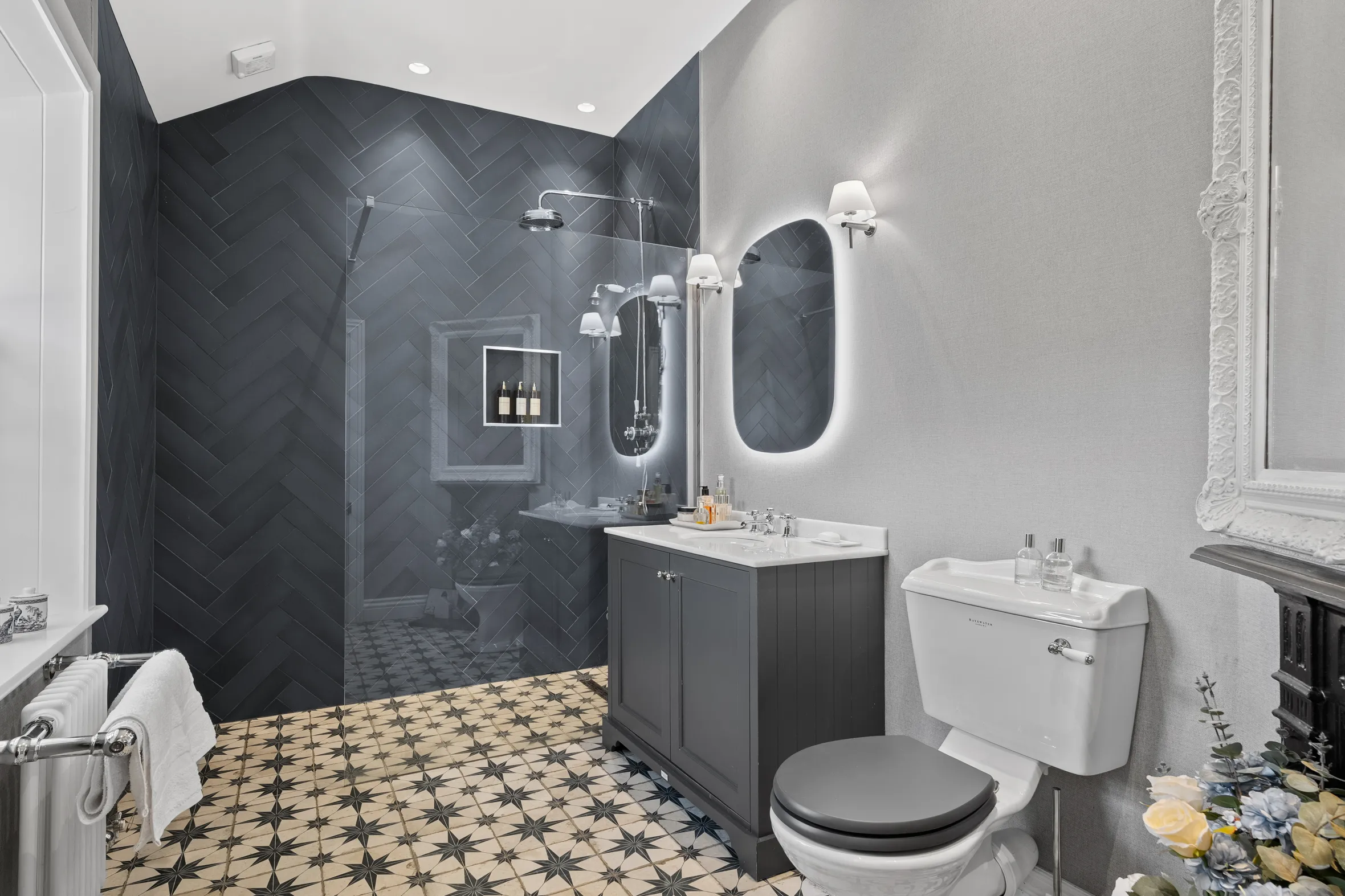 A bathroom with a walk-in shower featuring herringbone tile, a patterned floor, and a vanity with a backlit mirror.