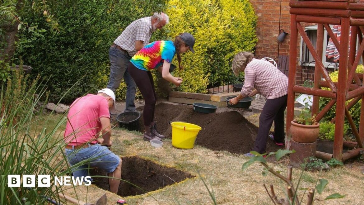 A view of four people digging a hole in a garden. One man in a pink t-shirt is inside a square hole, while three other people look through a pile of dirt.