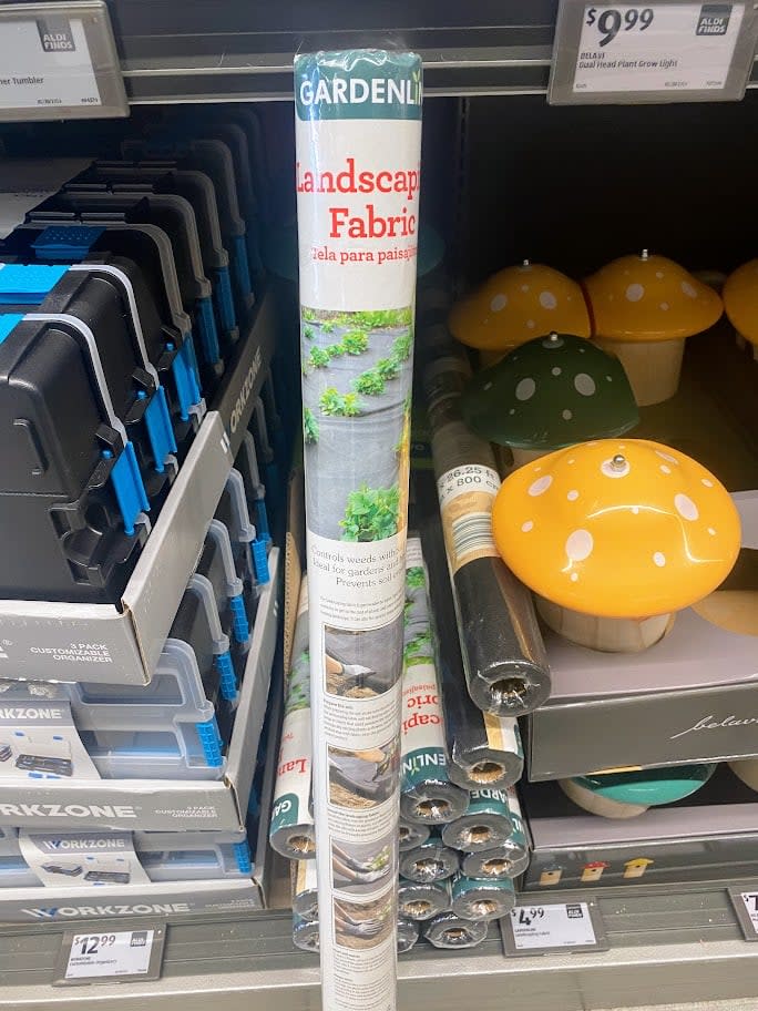 A roll of Gardenline landscape fabric is displayed on a store shelf between garden tools and decorative mushroom ornaments. The packaging shows images of fabric in use and includes product information. Price tags are visible above.