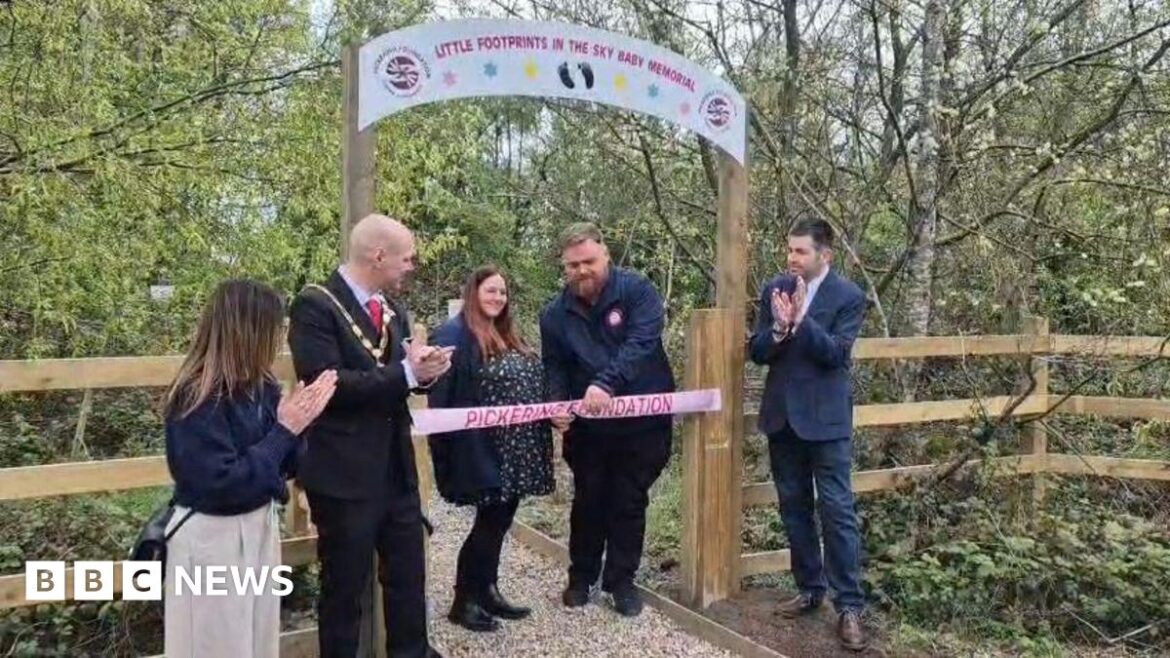 A man and a woman stand under a wooden archway in the middle of a wood, to cut a pink ribbon. Three other people next to them are clapping.