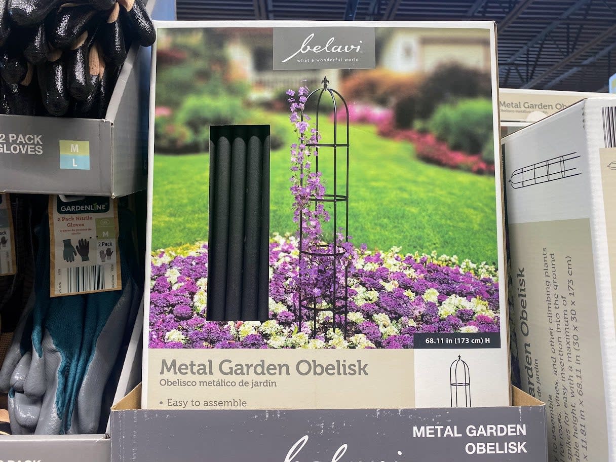 A box of a metal garden obelisk is displayed on a store shelf, showing an image of the product set in a garden with purple flowers and a climbing plant. The packaging highlights it is easy to assemble.