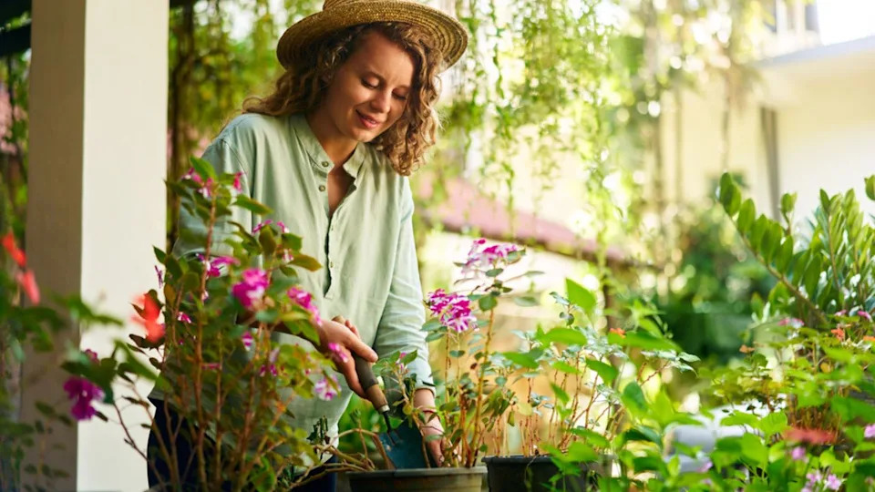 Young Woman Gardener in Straw Hat Holding Hand Shovel Taking Care of Potted Plants. Junior Caucasian Female Smiles Standing in Her Little Garden Planting Flowers in Pots.