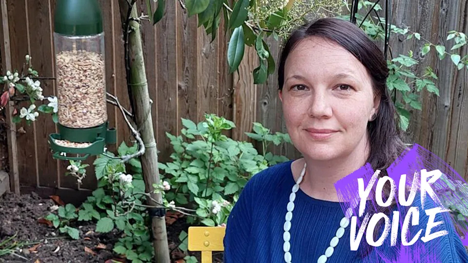 A woman with long brunette hair, wearing a blue jumper and a bead necklace stands in a garden next to a bird feeder. 