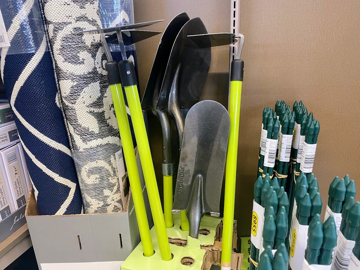 A display of gardening tools with bright yellow handles, including hoes and shovels, next to rolled rugs and bundles of green garden stakes in a store.