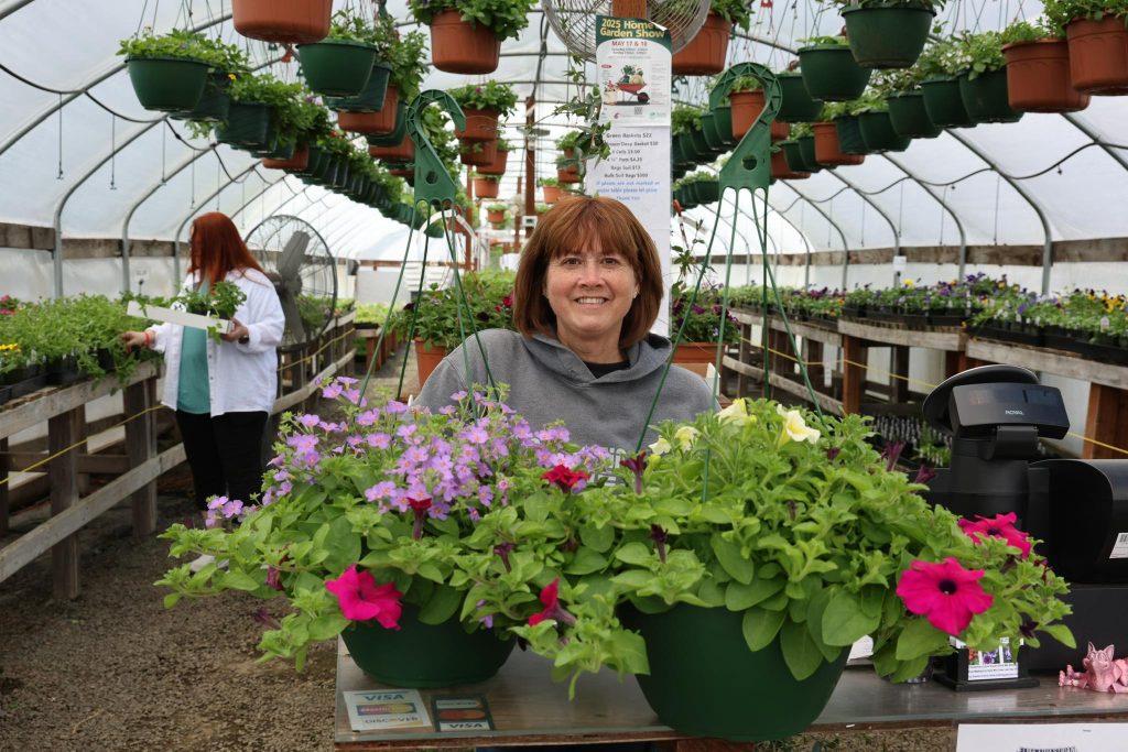 a woman stands behind two hanging baskets filled with flowers in a green house with rows of plants behind her