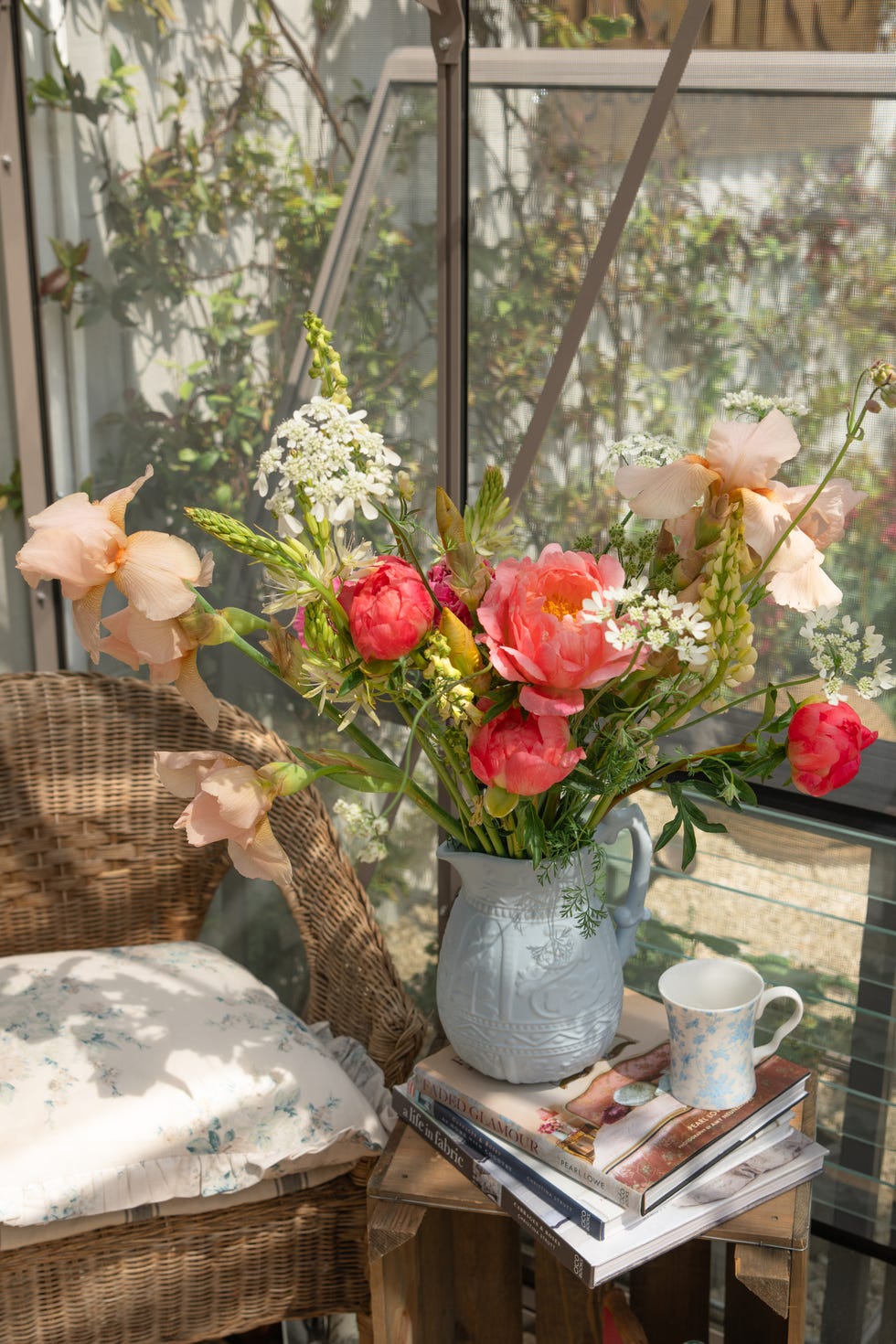 A bouquet of flowers in a ceramic pitcher on a wooden table next to a wicker chair.