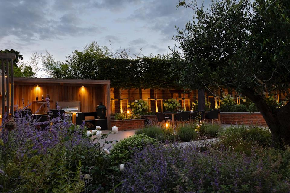 A garden at dusk with layered planting, an outdoor kitchen area, and uplights on a row of trees