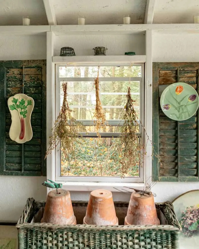 Woven basket with terracotta pots, dried herbs hanging by a window, and decorative plates on green shutters.