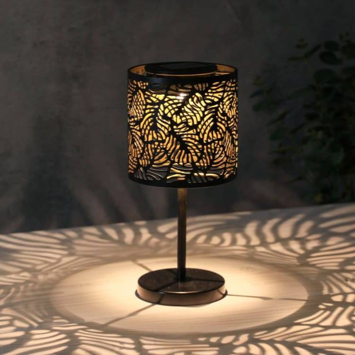 A table lamp with an intricately designed black shade adorned with a golden leaf pattern. The lamp is positioned on a flat surface, casting a warm, inviting glow and a delicate shadow pattern on the surrounding area.