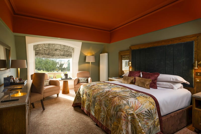Luxury hotel bedroom with tropical-print bedspread, terracotta ceiling, sage walls, and bay window garden view.