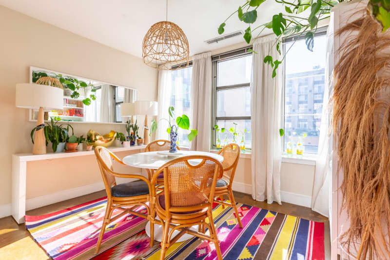 Dining room with round table, wicker chairs, colorful rug, plants, and large windows with sheer curtains.