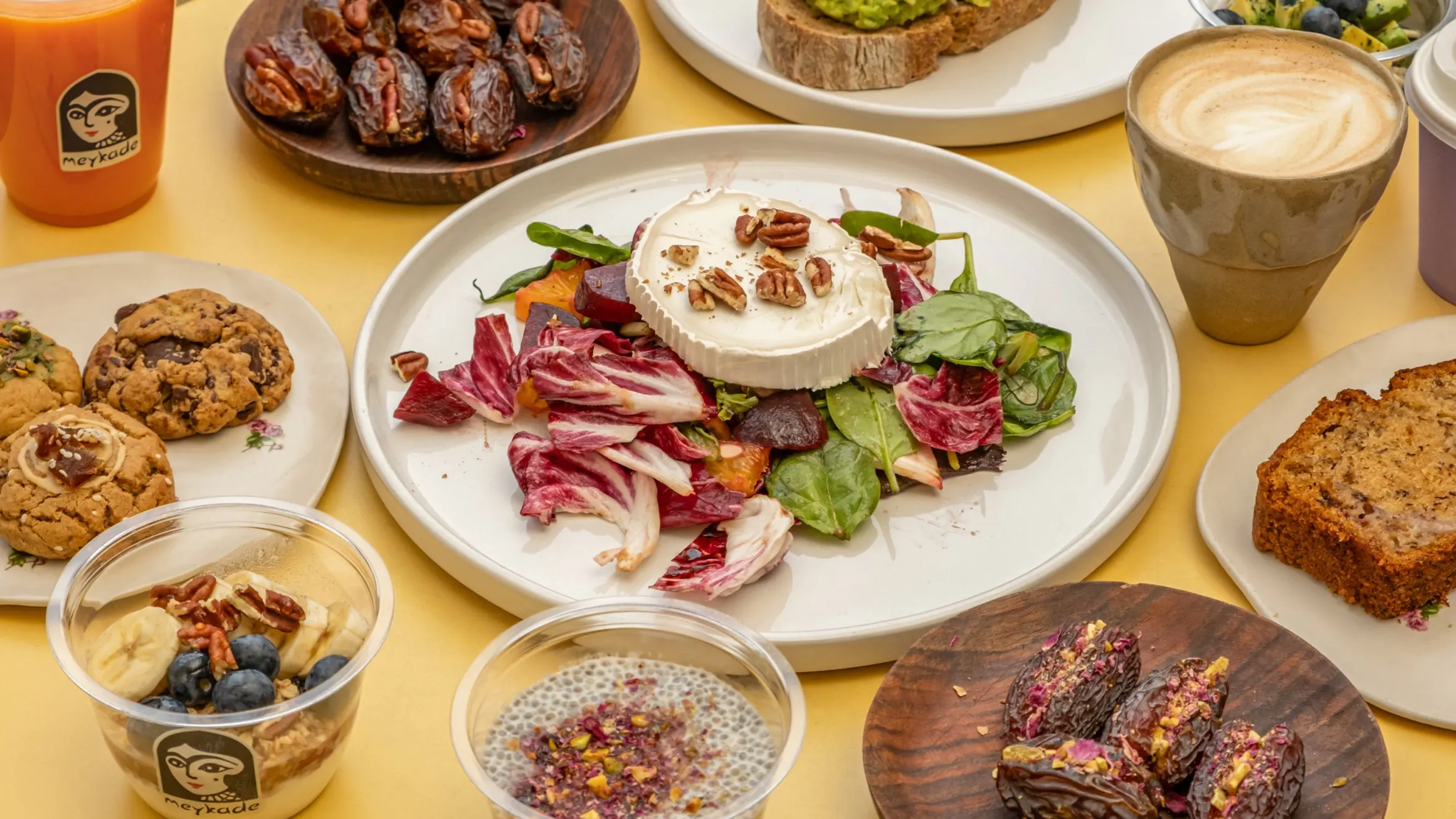 An array of food items from Meykade, including a salad with goat cheese, avocado toast, pastries, and drinks.