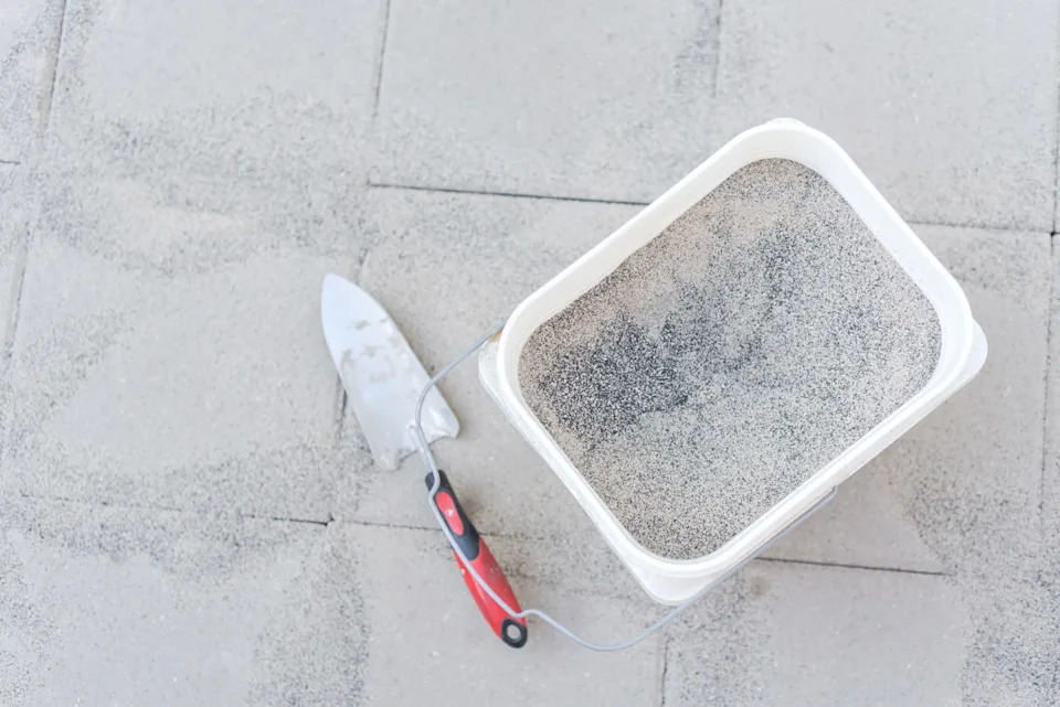 full bucket of polymeric paver sand with trowel on top of gray concrete patio stone paver walkway.