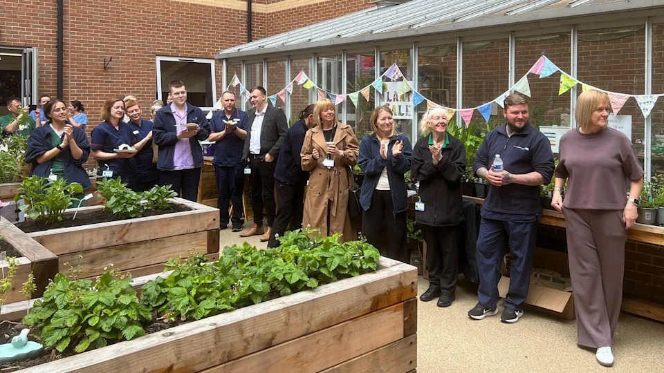 A group of people all stood in front of a greenhouse with bunting on it, they are clapping and smiling. Around them are raised flower beds.