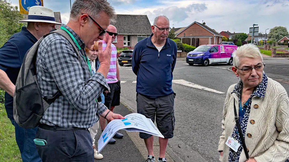 Six people are standing on different spots on a pavement. One is looking down at a map the others are looking to the right. Behind them is a roundabout and some bungalows