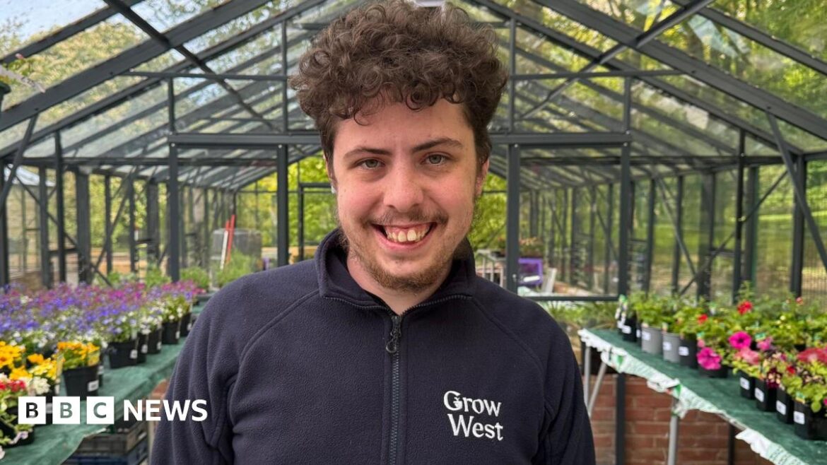 Workington gardens and cafe open to support community Ethan smiling into the camera while standing in one of the greenhouses. He has brown, curly hair and is wearing a blue zip-up fleece which reads 'Grow West' on the right of his chest. There are tables on either side of him lined with pots with different coloured flowers in them. On the right they are pink and on the left they are yellow, purple and pink.