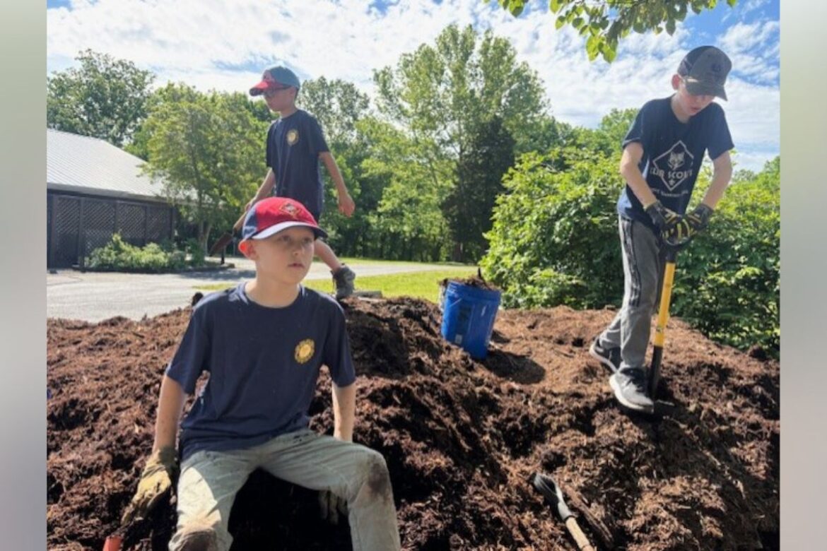 Three boys digging a large dirt mound outdoors on a sunny day, one seated while two stand with shovels nearby.