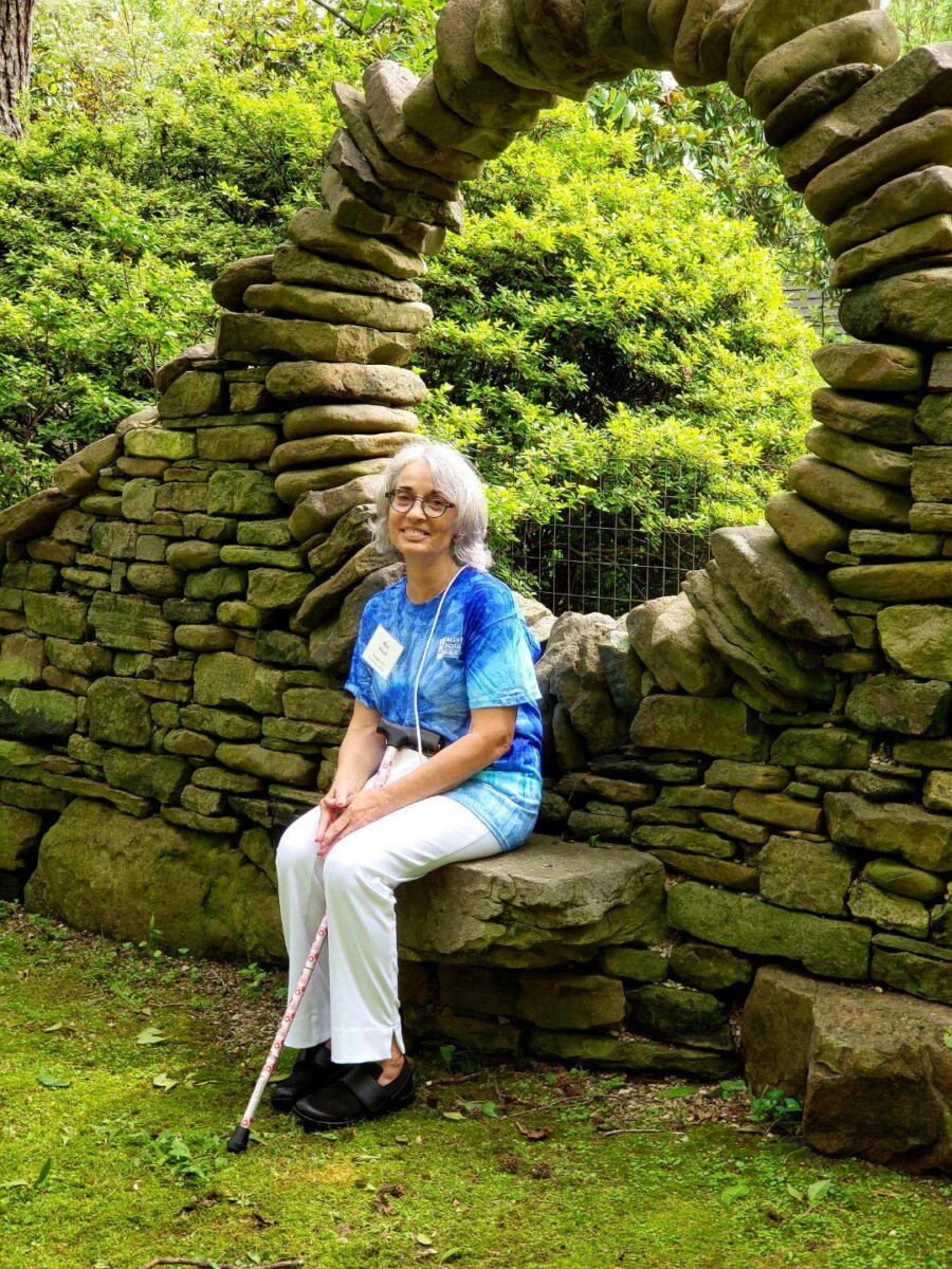 Meet the Memphis Gardener Who Sparked a Citywide Movement - 2 An older woman with gray hair, glasses, and a cane sits on a stone bench in front of a circular stone arch in a garden. She wears a blue shirt, white pants, and a name tag.