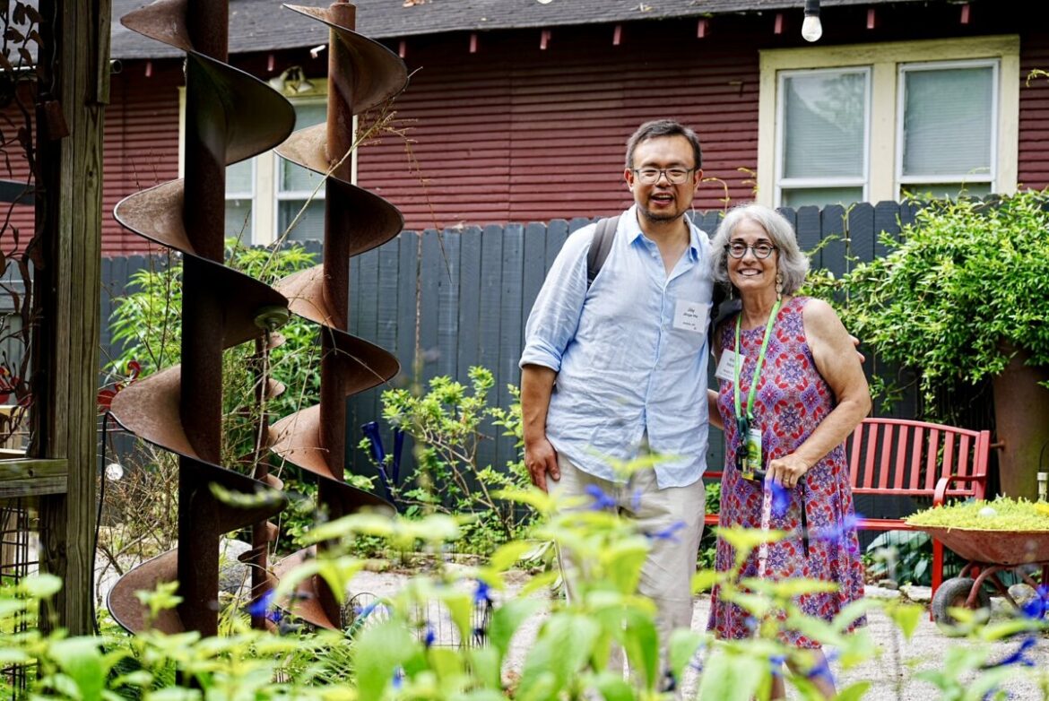 Meet Kim Halyak of Experience Memphis Gardens Two people stand together and smile in a lush garden, surrounded by plants, a large metal sculpture, and a red wooden bench. Created by Kim Halyak for FACES, the scene features a house and fence in the background.