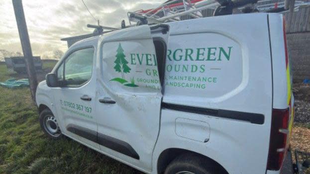 A white van parked up on grass with green letters saying Evergreen Grounds. The door is badly dented and partially hanging off.