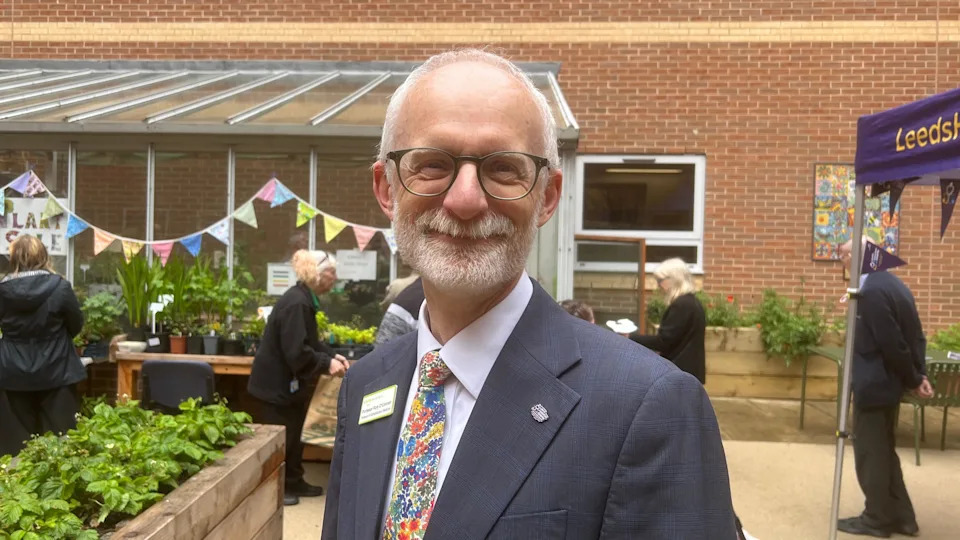 An older man with short white hair and beard is smiling at the camera. He has black round glasses and is wearing a suit jacket with a white shirt and floral tie. He is standing in front of flower beds and a table with potted plants on it. In the background there is a greenhouse with bunting hanging from it.