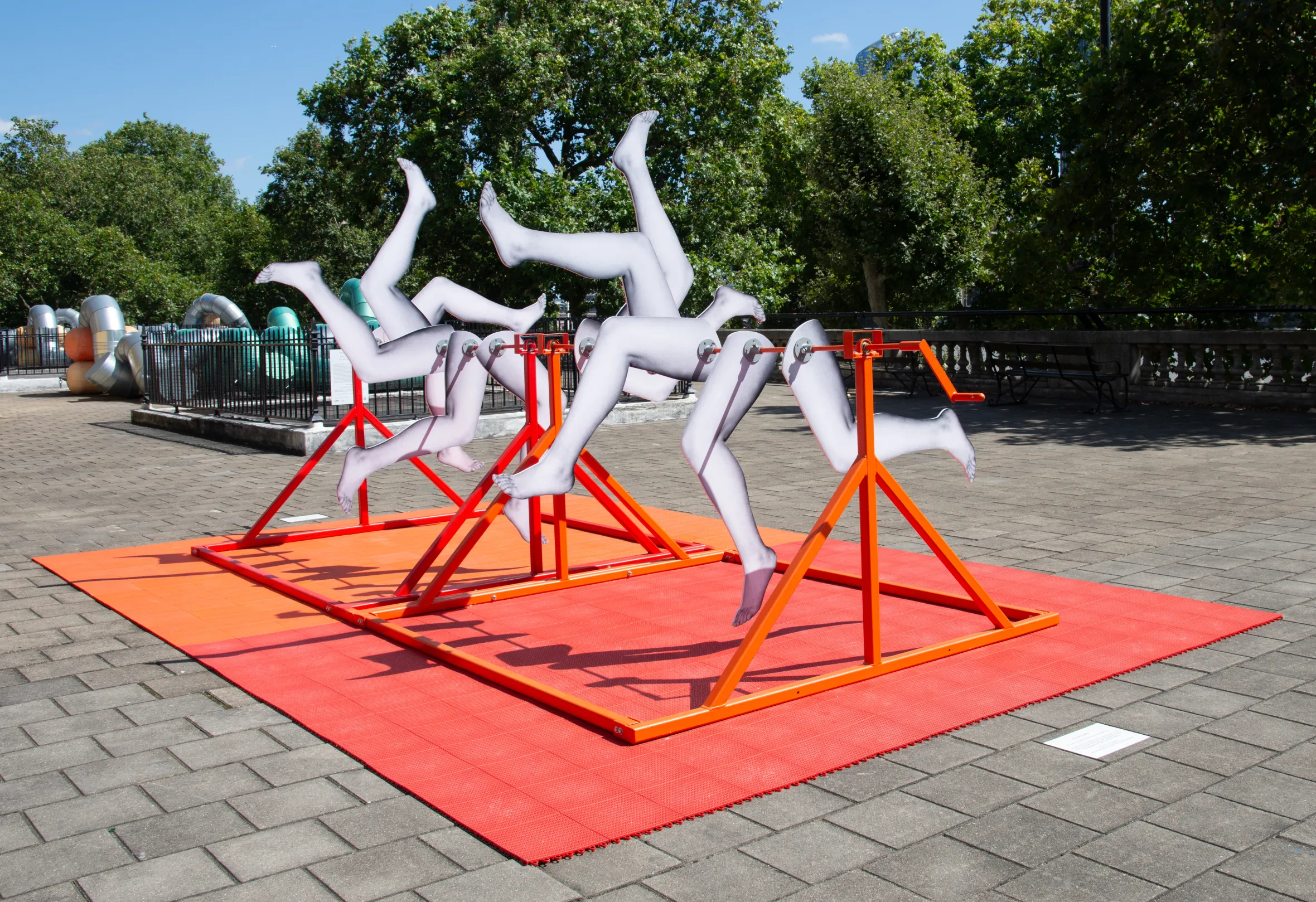 Kinetic art installation called "Its All Kicking Off" by Lucy Gregory featuring white cut-outs of legs mounted on orange frames on a red mat.