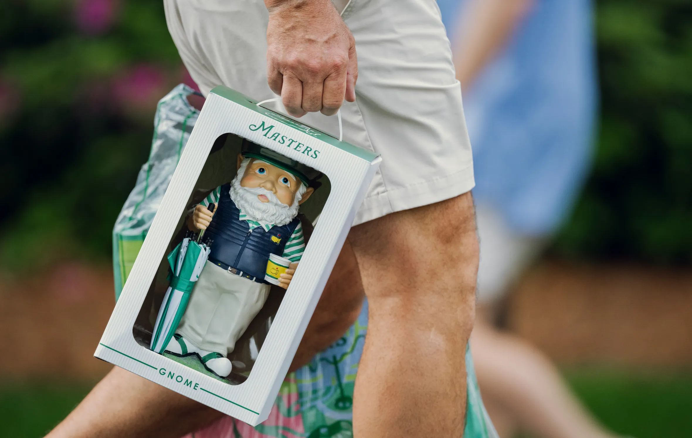 A person carries a Masters golf gnome in its box, holding a mini umbrella and a cup with the Masters logo.