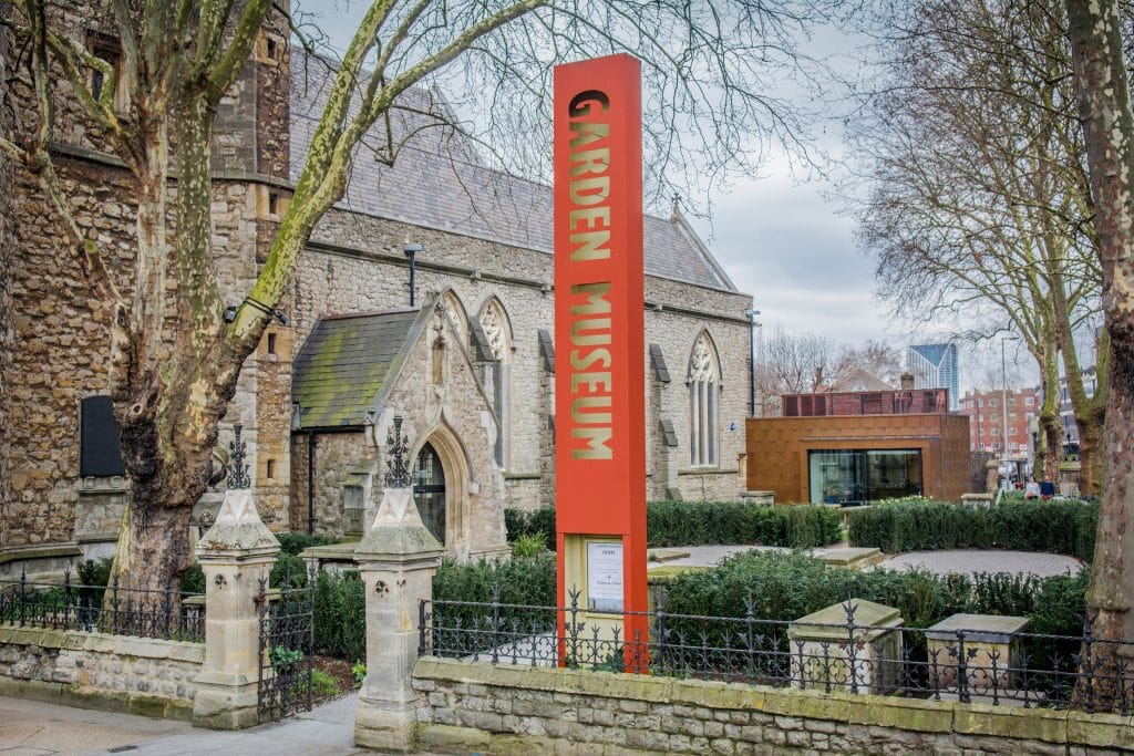 Red Garden Museum sign stands beside historic stone church building and leafless trees in London.