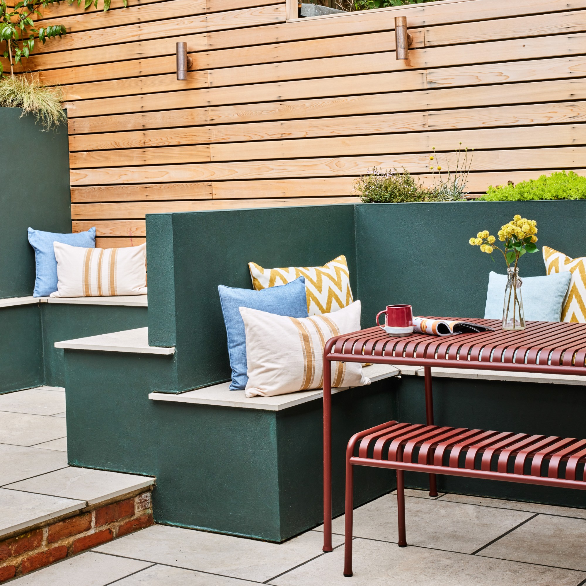 A garden with HAY's Palissade outdoor dining table and matching bench in red next to built-in bench seating painted dark green