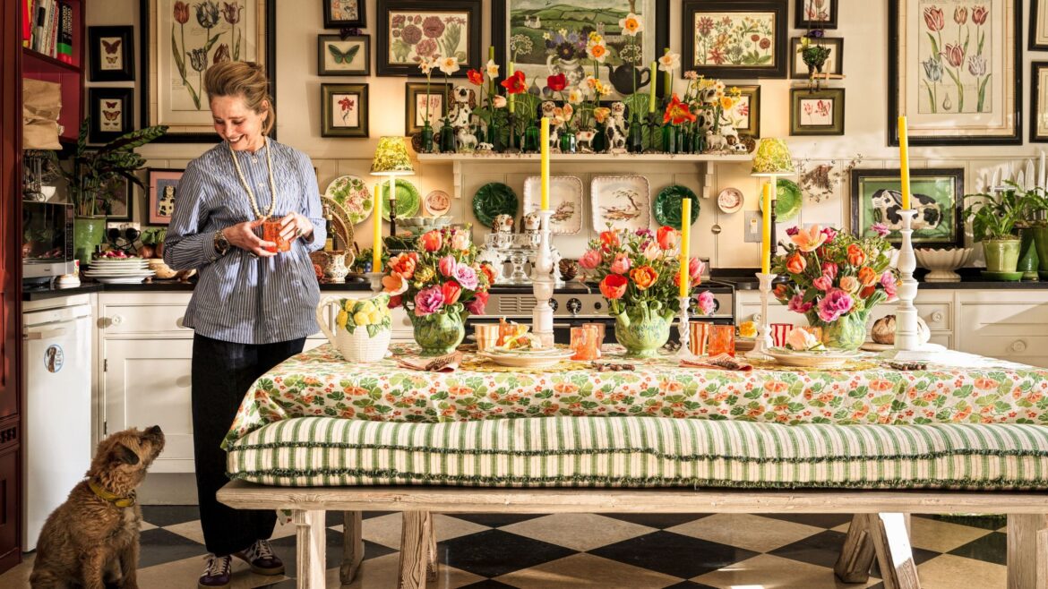 How I Host: A Floral-Inspired Spring Gathering With Butter Wakefield Butter Wakefield smiles in her maximalist kitchen, holding a drink next to a long dining table set with colorful floral arrangements, yellow candles, and a patterned tablecloth. A small terrier sits at her feet