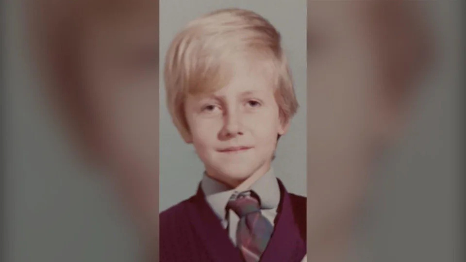Steve Damon is pictured as a child in the 1970s. The image is a school photograph and Steve is wearing a grey shirt, a dark coloured v-neck jumper and a green and burgundy school tie. He has floppy blonde hair. He looks sad in the picture.
