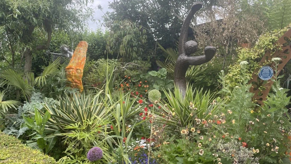 An exotic front garden can be seen. It is filled with sculptures, plants, flowers and trees.