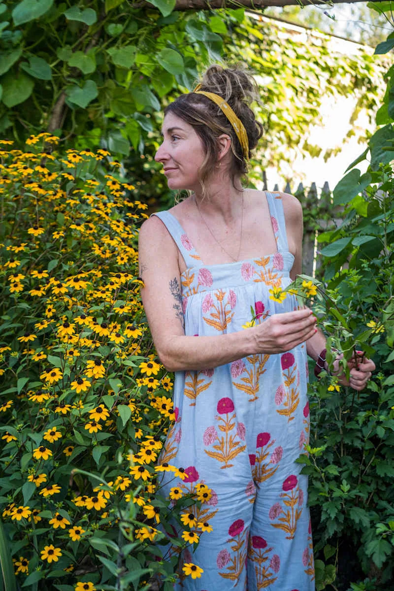Woman in a floral jumpsuit standing among vibrant yellow flowers, holding a branch in a lush garden.