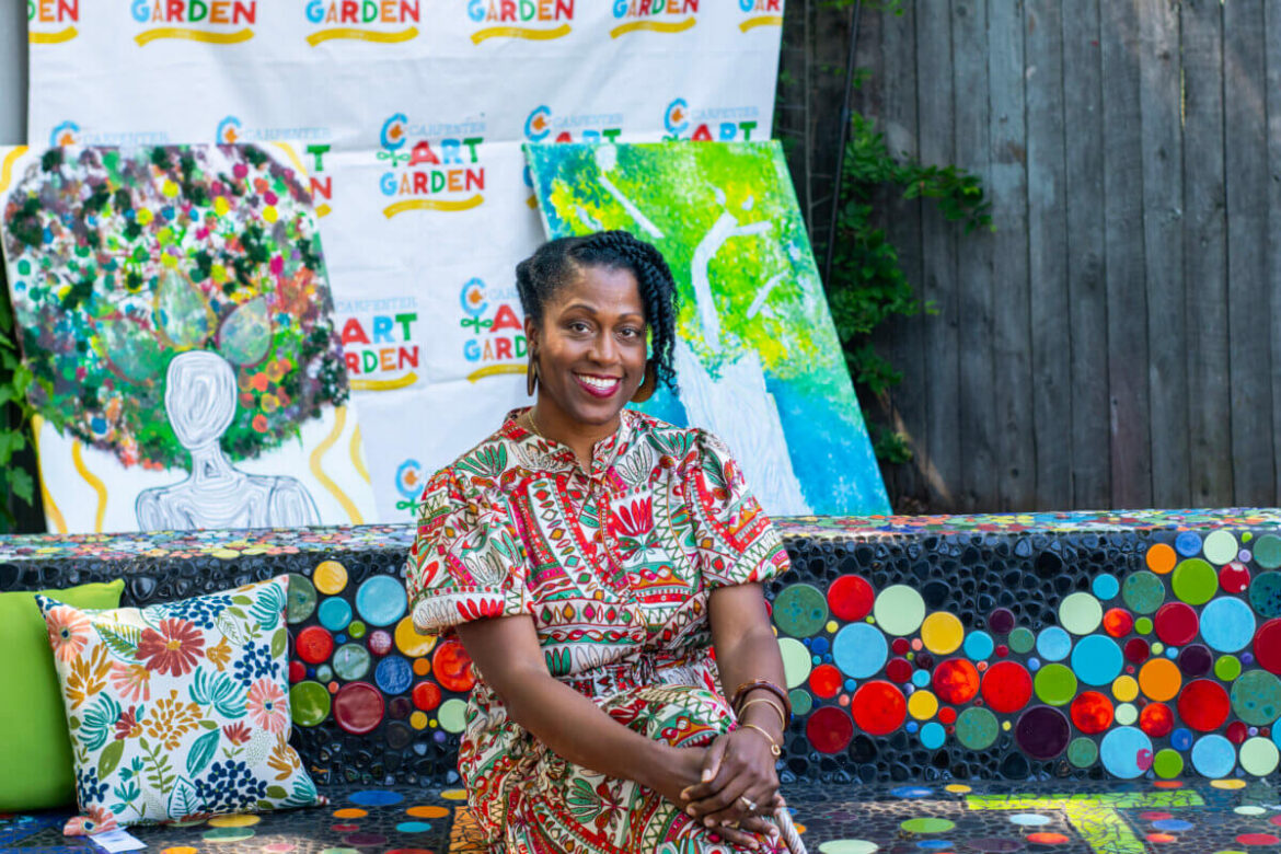 Meet Ayanna Murray of the Carpenter Art Garden A woman in a patterned dress sits on a colorful mosaic bench at the "Community Art Garden," with abstract paintings and a banner by Ayanna Murray in the background.