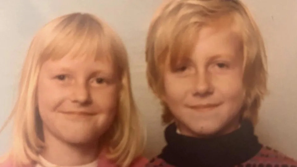 Steve Damon is pictured as a child sitting beside his sister in what looks like a school photograph from the 1970s. Both Steve and his sister have blonde hair. Steve is wearing a polo neck jumper and his sister is wearing a cream top with a pink cardigan over it. Both are smiling though Steve's smile looks a little sad.