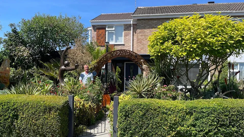 Steve Damon is pictured standing in his front garden. He is surrounded by tall trees and shrubbery and there is a bronze statue of a figure with arms outstretched beside him. There is a rusted bronze arch on an angle on the other side of him. The arch is over a pathway which leads to his front door. A neat front hedge and a black metal gate can be seen in front of the garden.