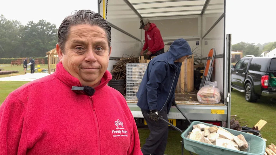 A man in a red fleece grimaces at the pouring rain while a colleague, hood up against the weather, pushes a wheelbarrow full of decorative stone.