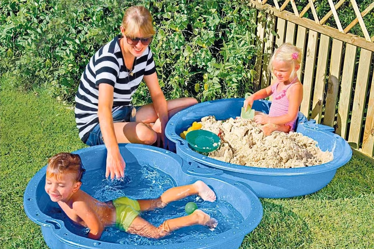 Two children playing in a sand and water pit while a woman sits nearby 