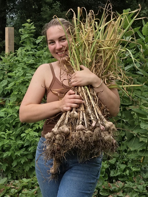 A smiling woman holds a large bundle of dried plants in a lush garden setting.