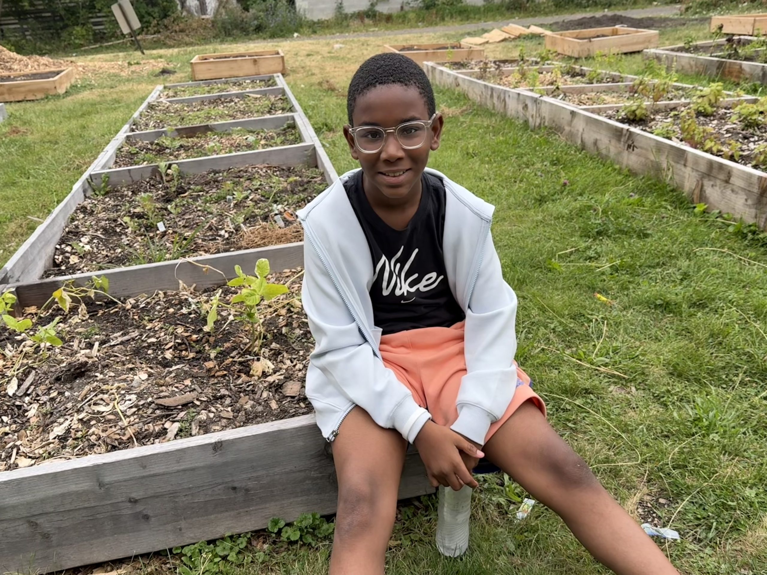 A person wearing glasses and a white jacket over a black Nike T-shirt and holding a water bottle sits on the edge of a raised garden bed with small plants, surrounded by additional wooden garden beds and grass.