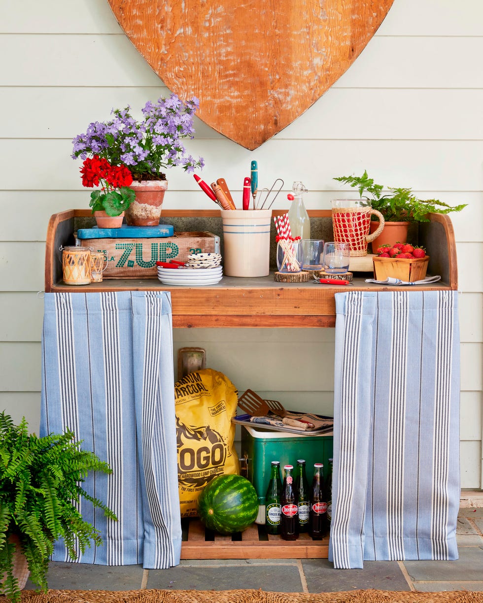 Wooden table decorated with flowers, kitchen utensils, and drinks, featuring blue striped curtains and a watermelon. Wooden table decorated with flowers, kitchen utensils, and drinks, featuring blue striped curtains and a watermelon.