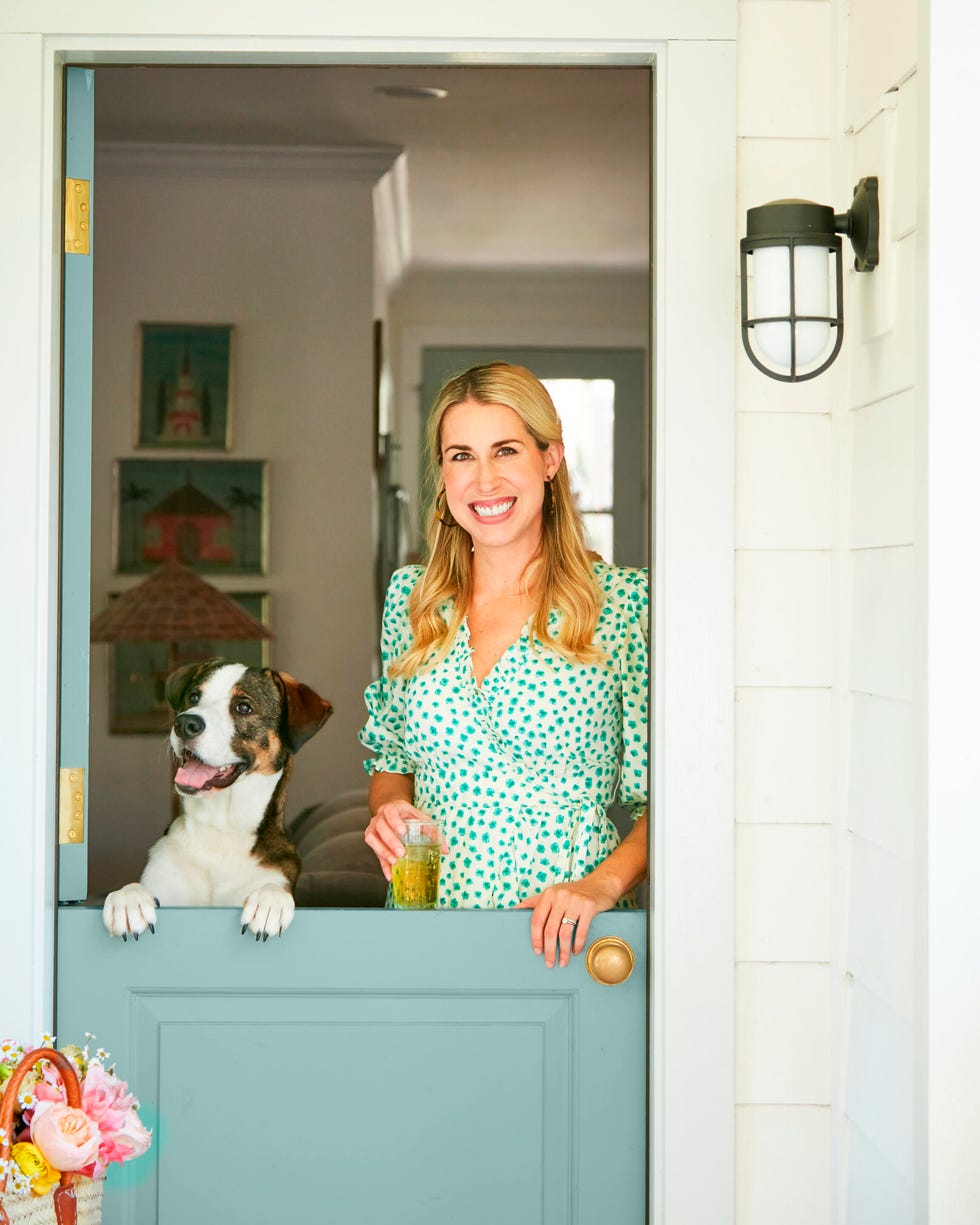 A woman with a drink smiles beside a dog at a doorway, surrounded by a light and welcoming home atmosphere.
