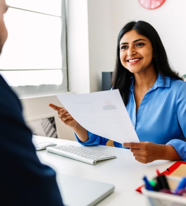 Confident successful Indian female HR recruitment manager holding resume in hands while having an interview in a modern office, looking and listening to job candidate. Friendly interview between HR female director and caucasian job candidate. - stock photo