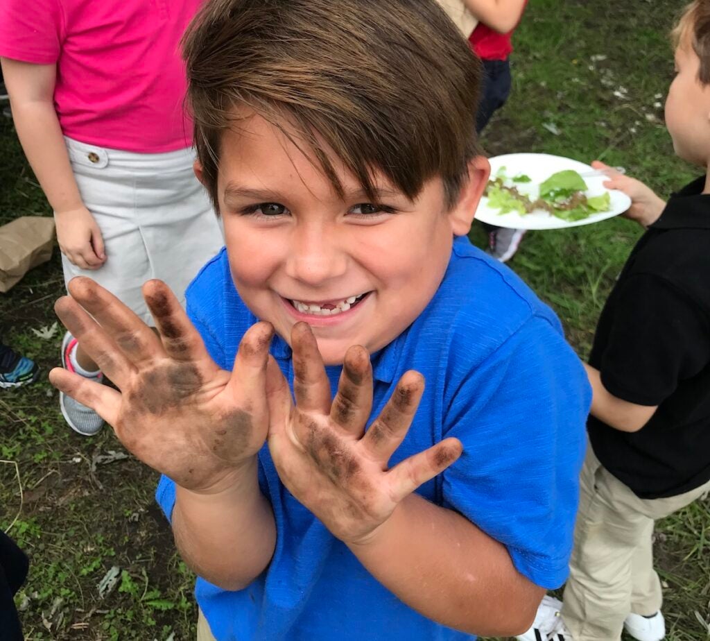 A student holds up his hands covered with dirt and smiles.