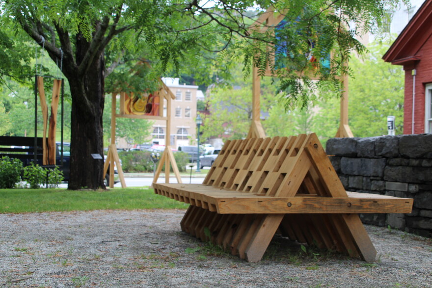 A wooden bench sits in a sculpture garden. There is a tree to the left and wooden sculptures behind the bench and tree.