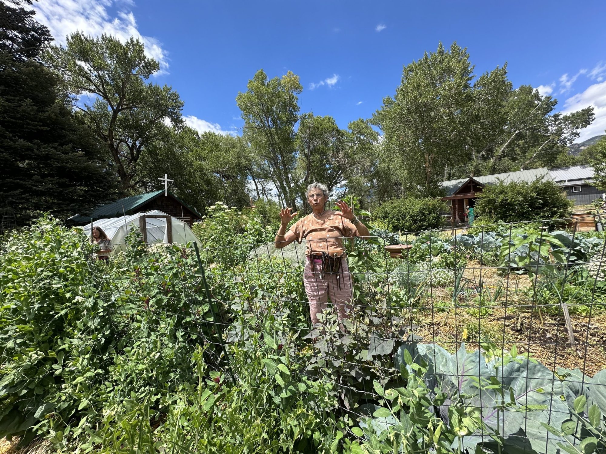 A woman wearing an orange shirt and tan pants with a tool belt around her waist gestures with her hands in a large garden that is helping to resolve Crestone's "food desert" problem