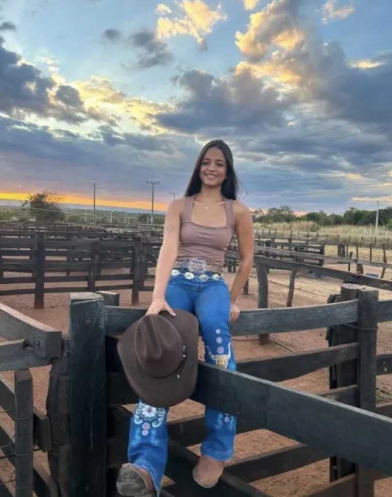 Ana Beatriz Araújo Ferreira sitting on a wooden fence, holding a cowboy hat, with a sunset sky in the background.